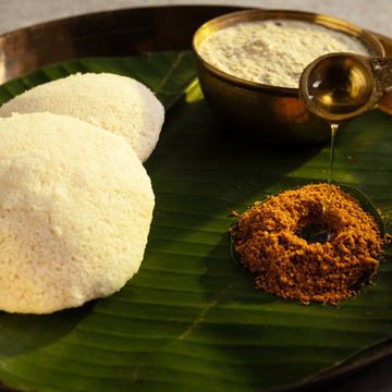Two white idlis on a banana leaf with chutney and sambar on a metal tray.
